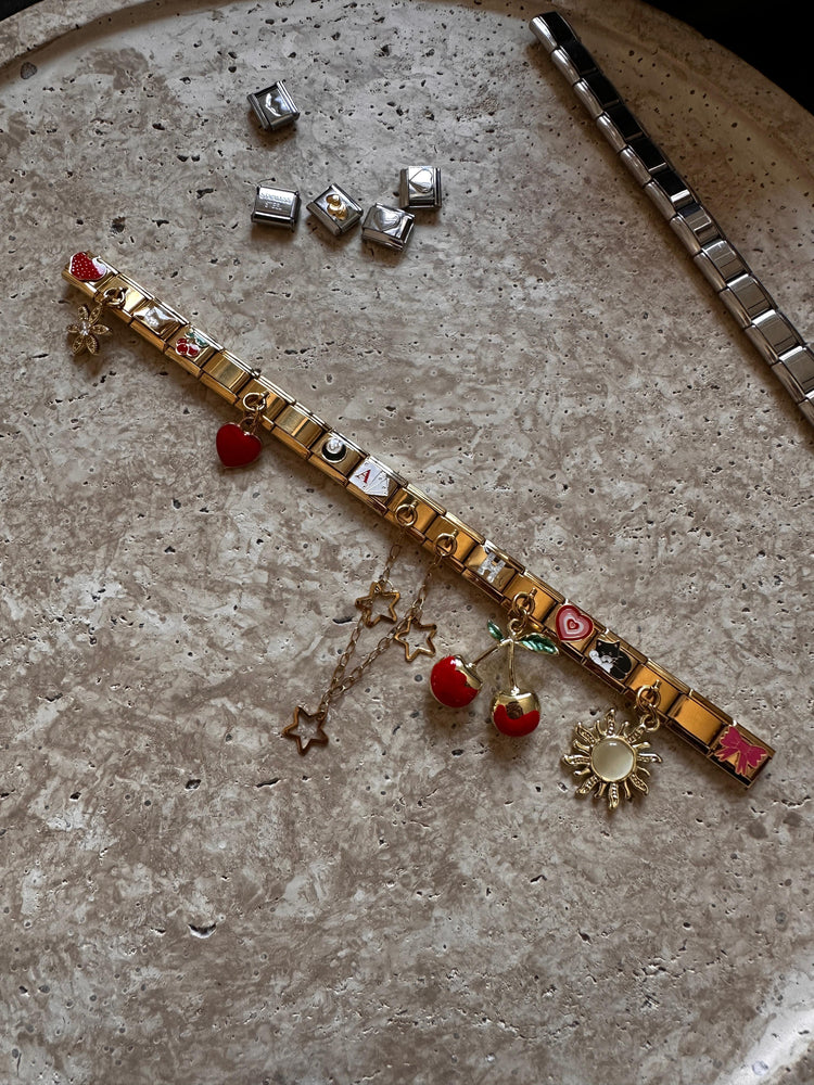 Goldenes Italian Charm Bracelet mit verschiedenen Charms, darunter Kirschen, Sonne, Sterne und Herz, flach auf einer strukturierten Steinoberfläche arrangiert.